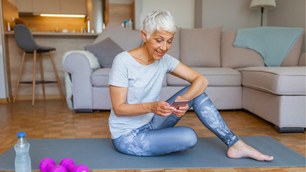 Ältere Dame in Sportkleidung auf Gymnastikmatte mit Handy in der Hand
